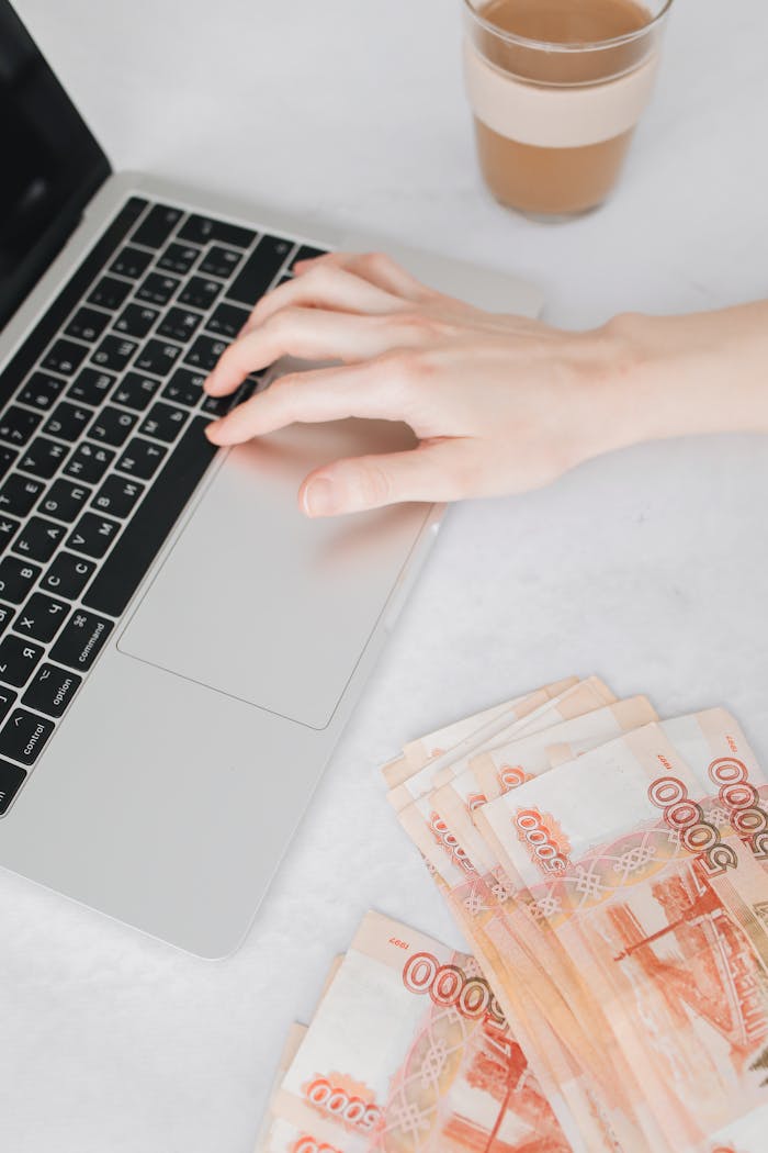 A person typing on a laptop next to a stack of Russian Rubles on a white table.